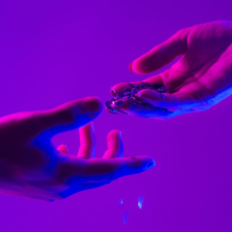 Dettaglio di due mani che si sfiorano scambiando piccoli frammenti sotto luci neon, simbolo di contatto e tecnologia